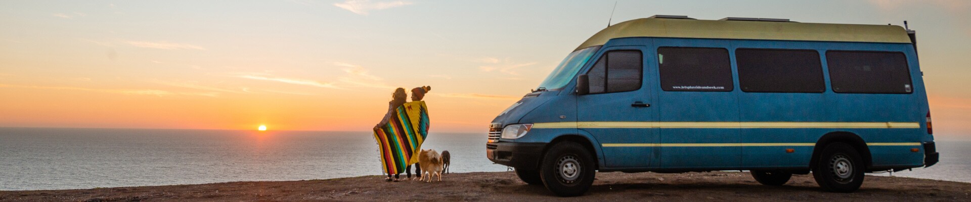 A couple wrapped in a brightly colored blanket stand outdoors next to a blue campervan. Two small dogs are at their feet.