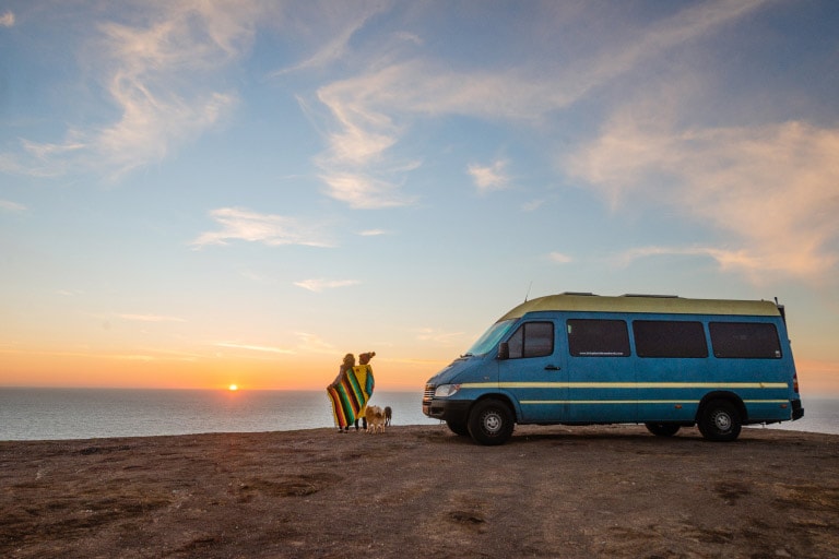 A couple wrapped in a brightly colored blanket stand outdoors next to a blue campervan. Two small dogs are at their feet.