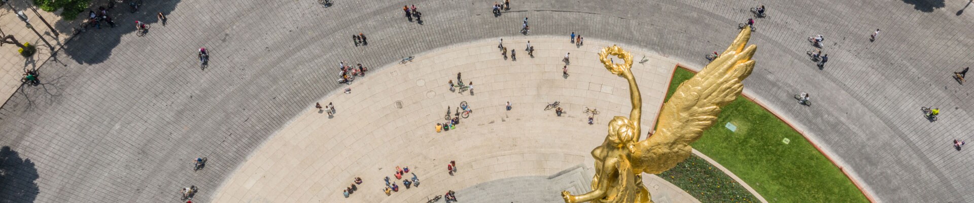 Aerial view of the golden Independence Monument in Mexico City. People are sitting, walking, or cycling around its base.