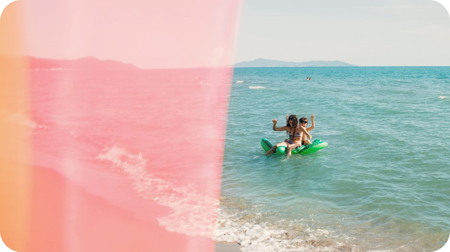 Dos personas en el océano sobre un inflable verde en forma de tortuga. Están sonriendo y alzando las manos.