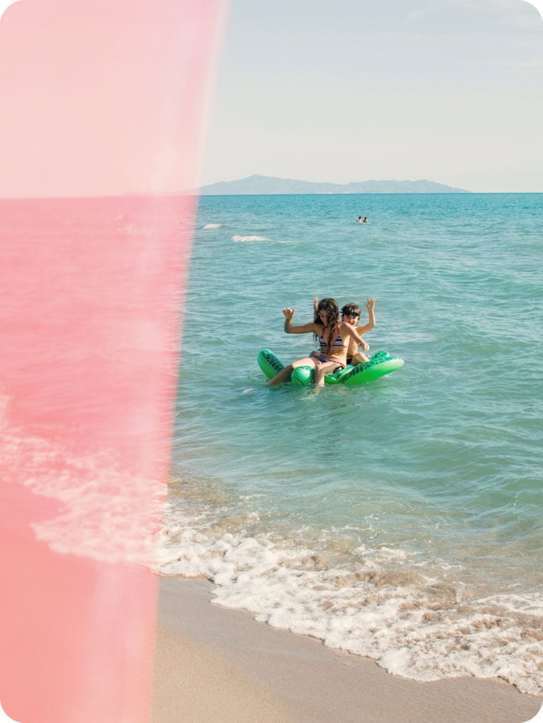 Dos personas en el océano sobre un inflable verde en forma de tortuga. Están sonriendo y alzando las manos.