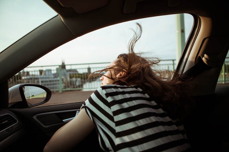 A person in a black-and-white striped shirt leans out a car window as the wind blows their long brown hair around.