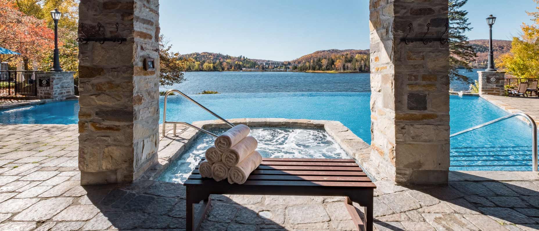 Rolled-up towels on a bench sit beside a bubbling hot tub connected to a blue shimmering infinity pool, overlooking a beautiful lake surrounded by mountains and trees. 