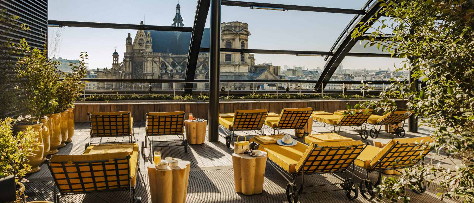 Elegant rooftop deck with huge windows looking out over the city, filled with mustard yellow loungers and serene foliage. 