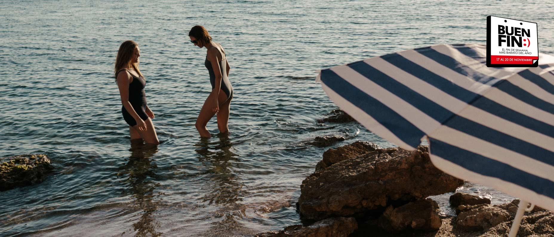 Dos personas están dentro de un lago y el agua les llega a las rodillas. A la derecha se ven piedras grandes y una sombrilla.