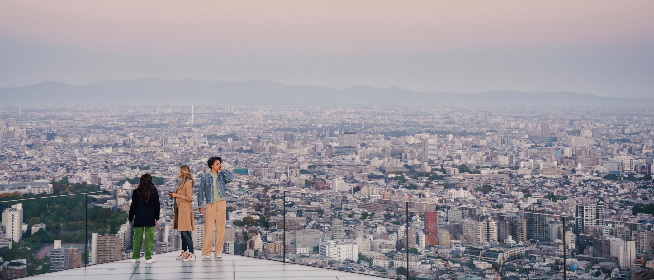 Three people stand on a skyscraper patio with clear walls in Tokyo. City buildings and mountains are visible behind them.