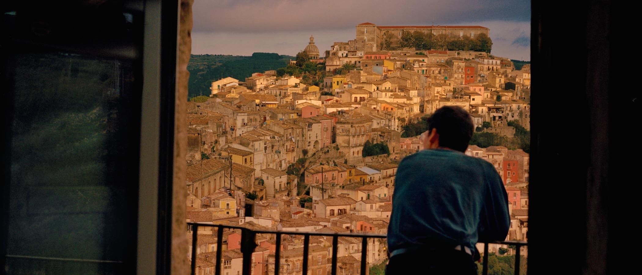 A person leans on a balcony railing and faces a hillside covered with antique buildings in Sicily.