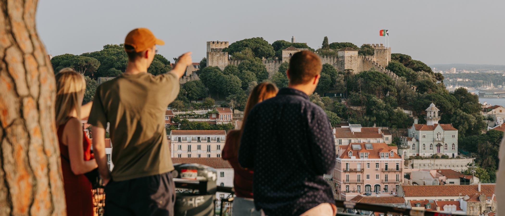 Four people stand on a balcony overlooking Lisbon. Two of the people point at a castle in the distance.