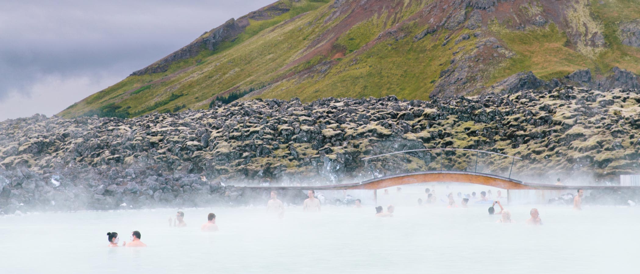 Several people swim in Reykjavik's Blue Lagoon as steam rises from the water. Rocks and a grassy hill surround the lagoon.