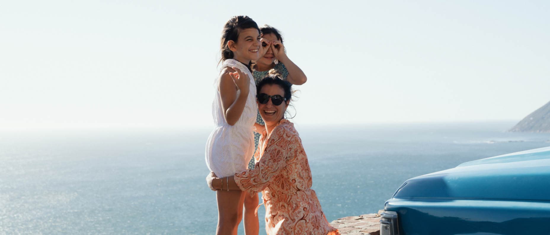 A smiling woman kneels and embraces two small girls while smiling at the camera. In the distance we see vast ocean and horizon witha bit of hilly coastline. The front of a blue car extends into the frame.