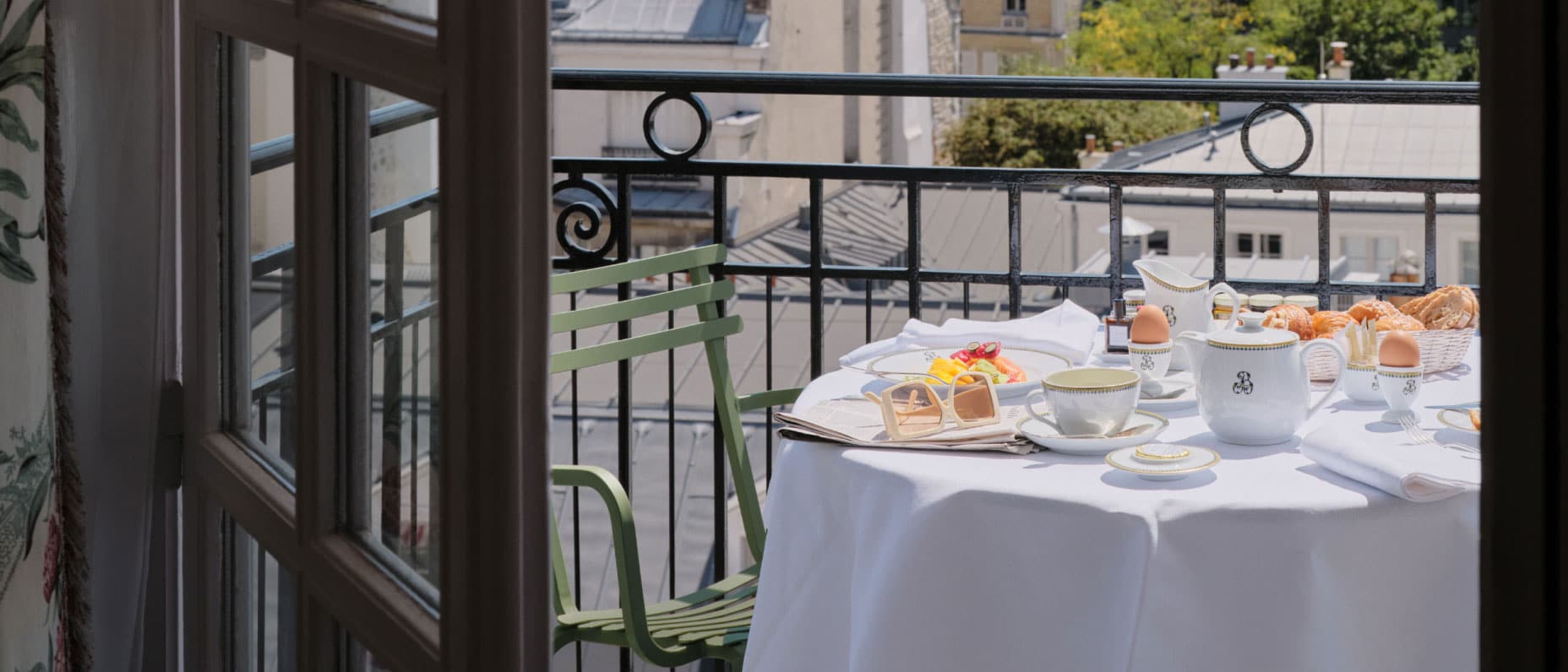 A pair of open French doors opens onto a small balcony with city views and a table set for two featuring a breakfast spread.