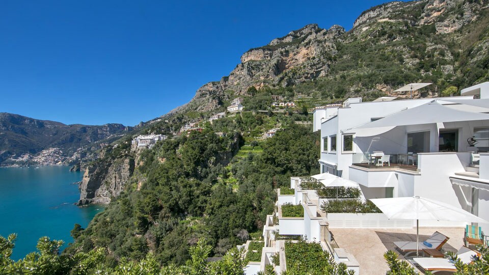 The white angular buildings of Casa Angelina in Praiano Italy rest against tree-covered cliffs by the Amalfi Coast.