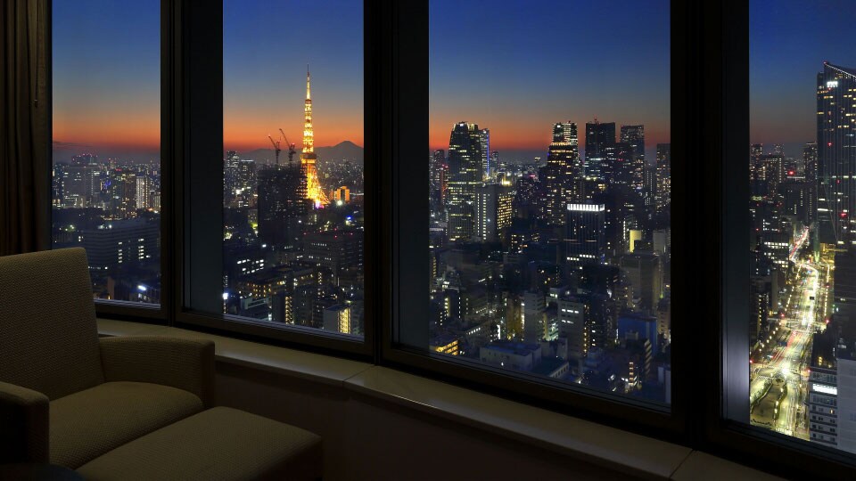 An upholstered chair and ottoman sit by a multi-panel window showing the Tokyo skyline at dusk at the Park Hotel Tokyo.