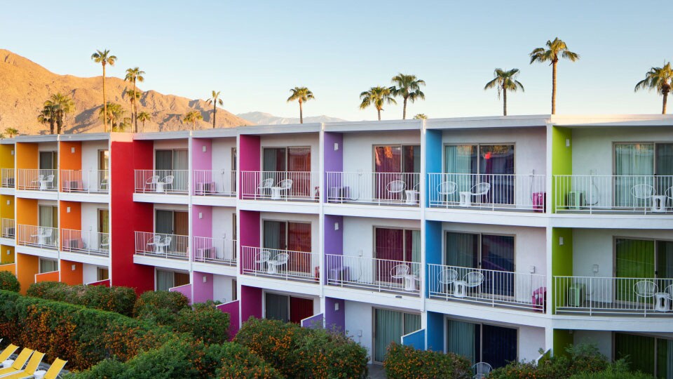 At the Saguaro Palm Springs a white hotel with rainbow-coloured accent walls has palm trees and mountains behind it.