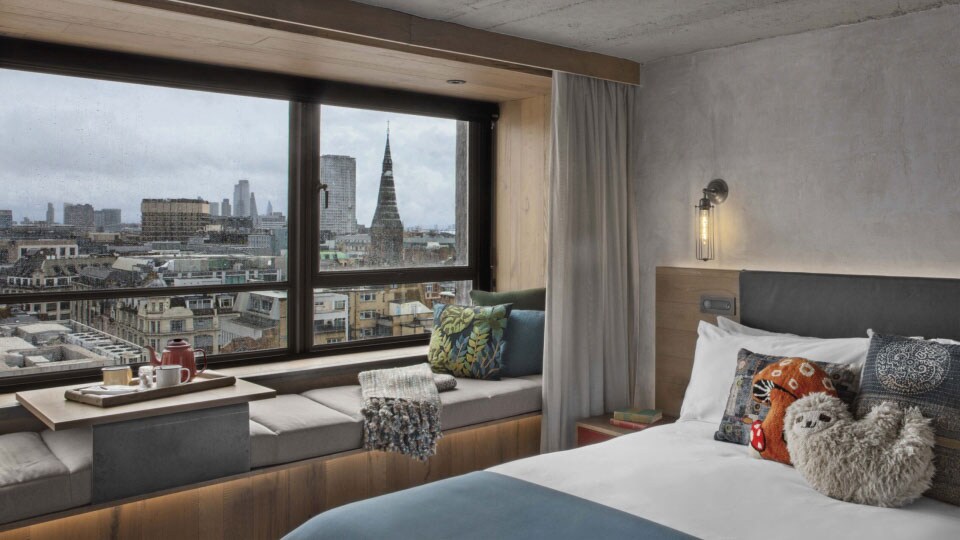 A Treehouse Hotel London room with a blue and white bed a gray built-in seating area beside a window and a city view.