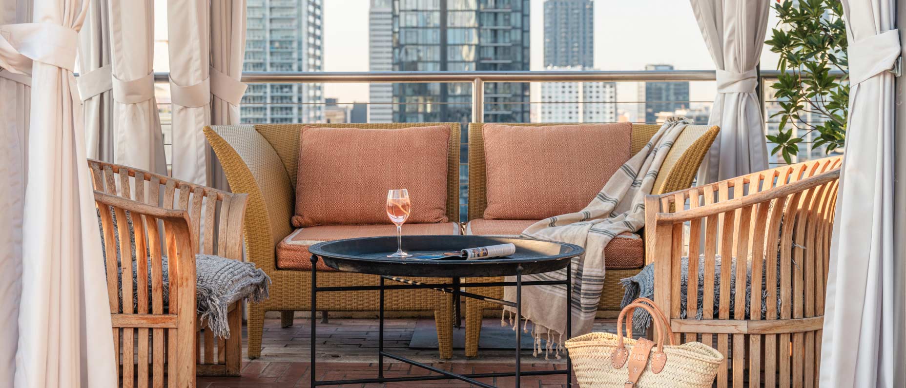 Un balcon au mobilier en rotin surplombe la ville. Un magazine et un verre de rosé sont posés sur la table basse.