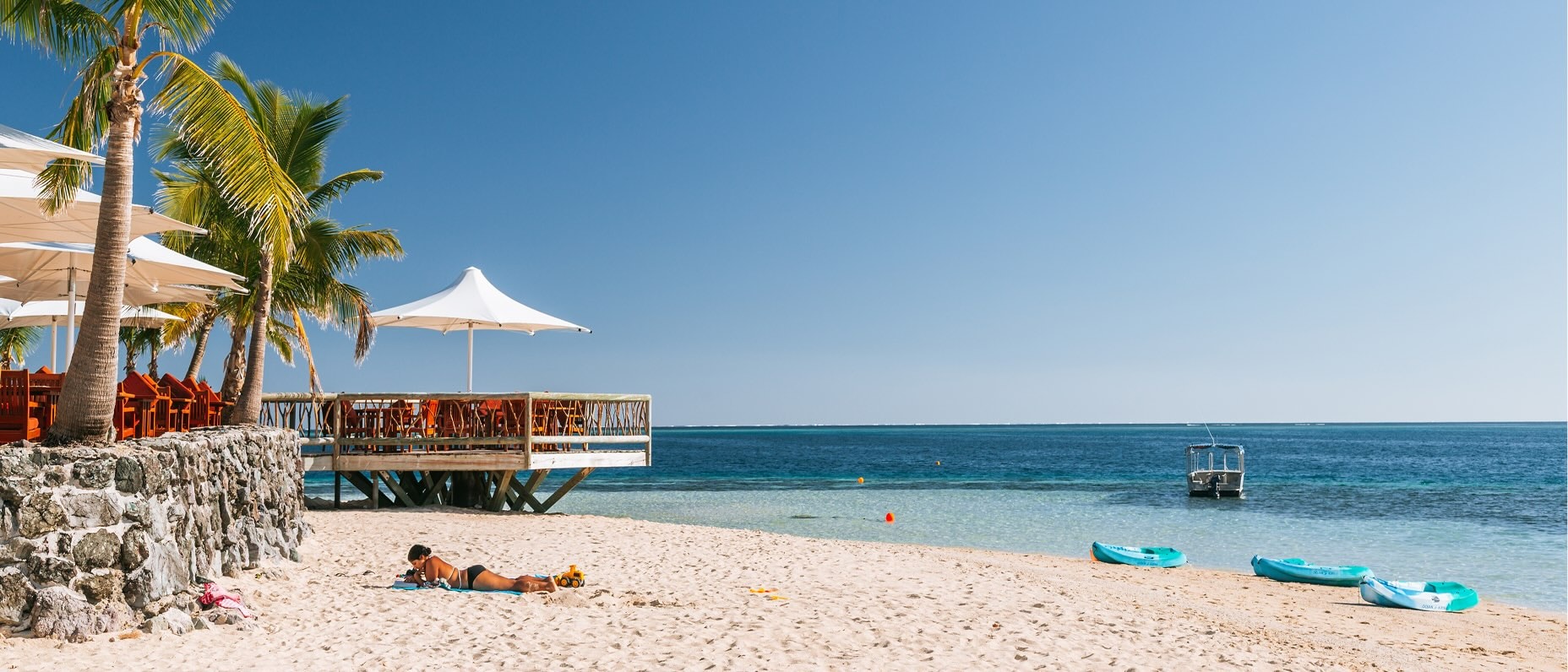 Sunny tropical beach with palm trees, a waterfront bar deck and kayaks on the sand beside calm turquoise water.
