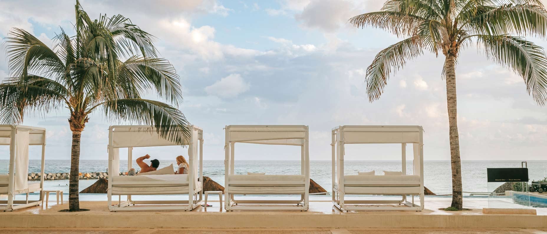 Three elegant beachside beds with white drapes in front of a swimming pool, overlooking the ocean.