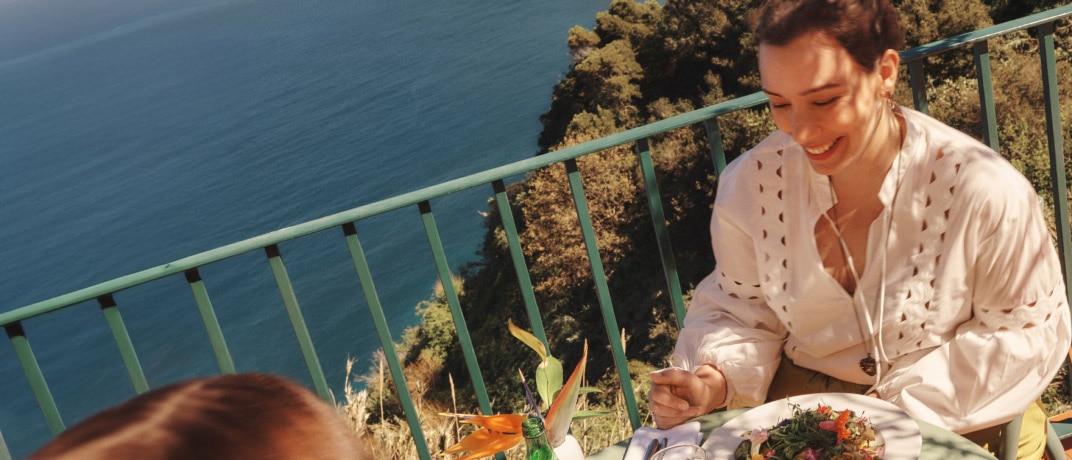 Two people sit opposite each other eating a meal with mountains and the ocean in the background.