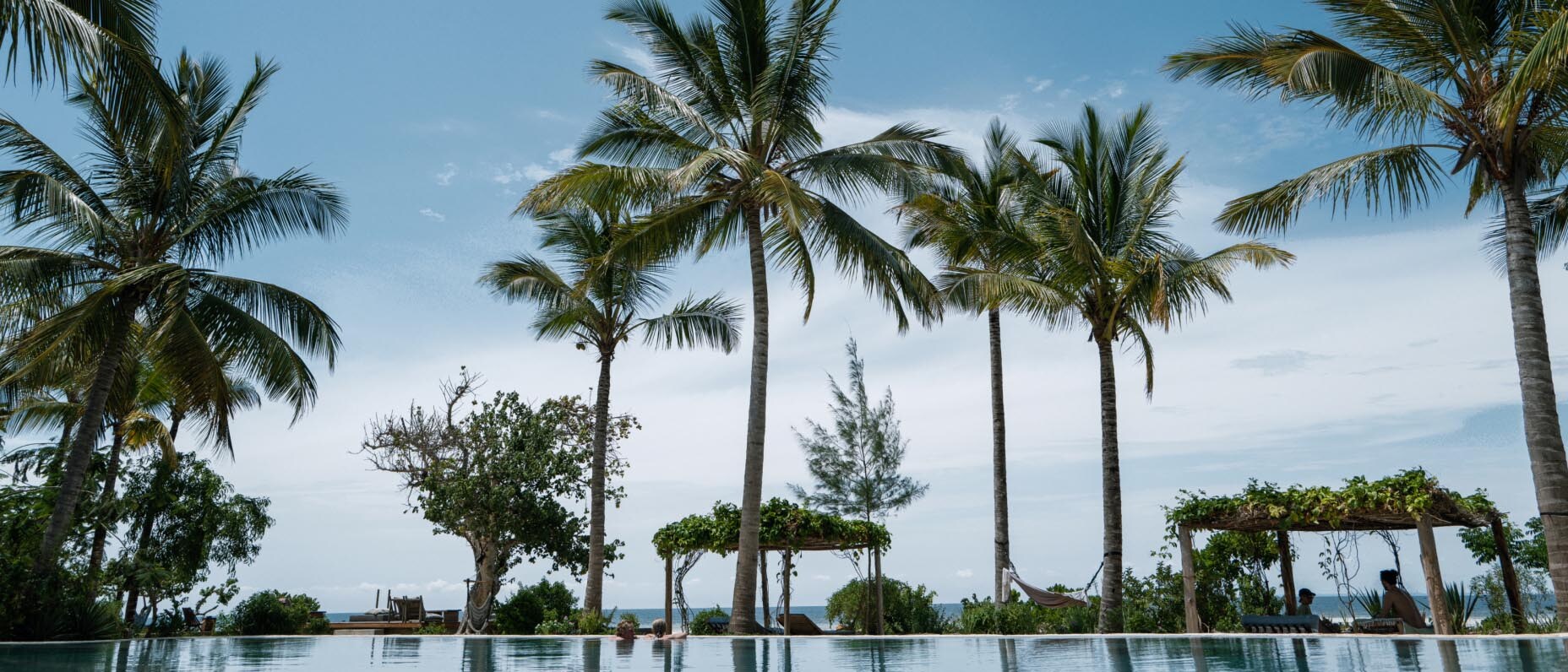 真っ青な空の下、インフィニティプールと海の境目にヤシの木が並んで見え、2 人の旅行者がプールの縁にもたれかかっている。