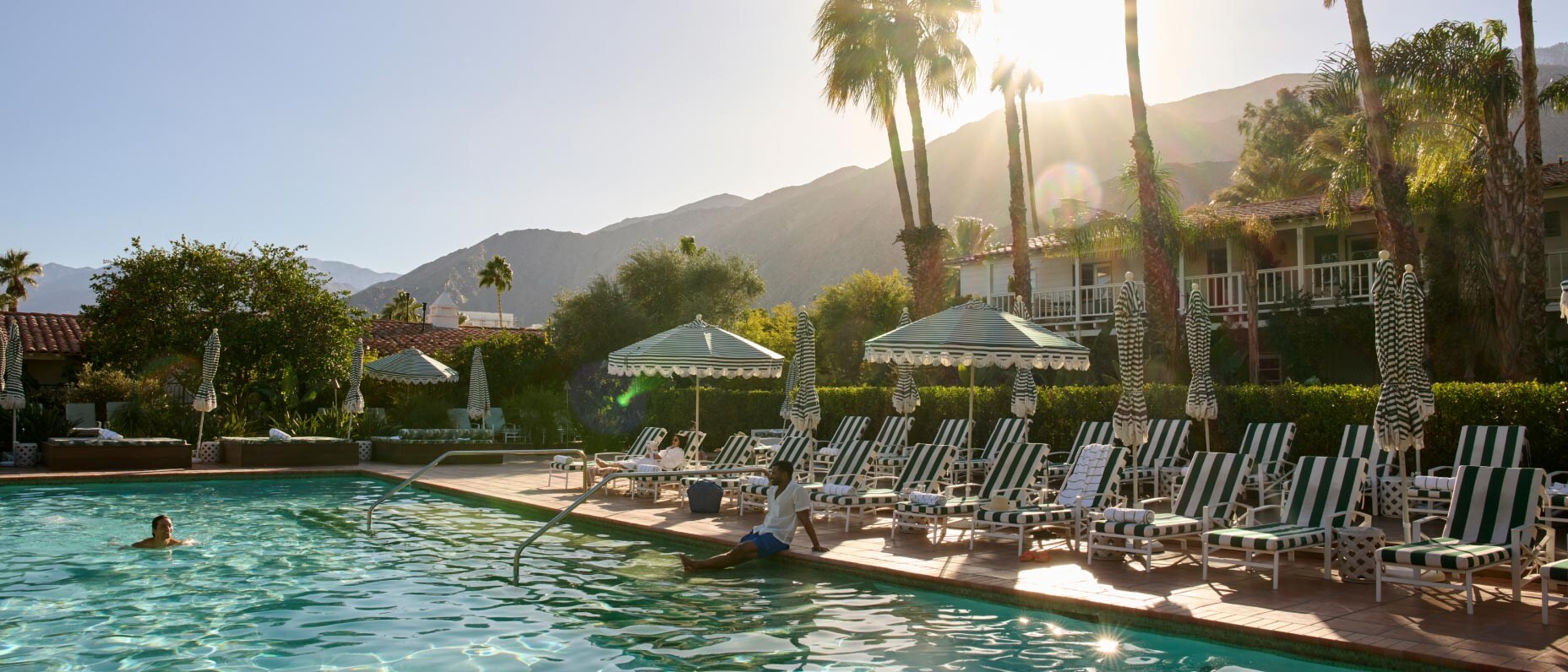 Outdoor pool with striped loungers and umbrellas, framed by palm trees and mountain views in the evening sun.