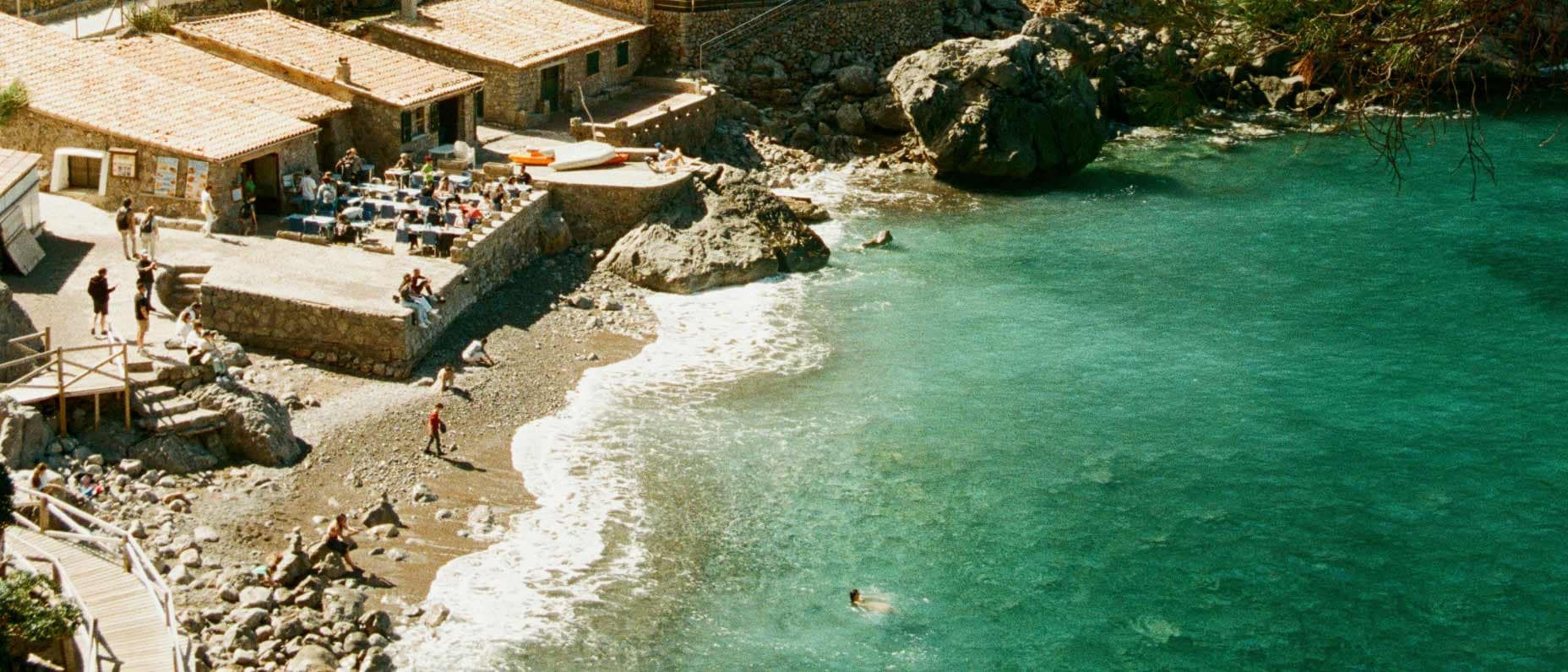 In a secluded rocky cove, visitors dine at an outdoor restaurant, relax on the beach and swim in the blue-green sea.