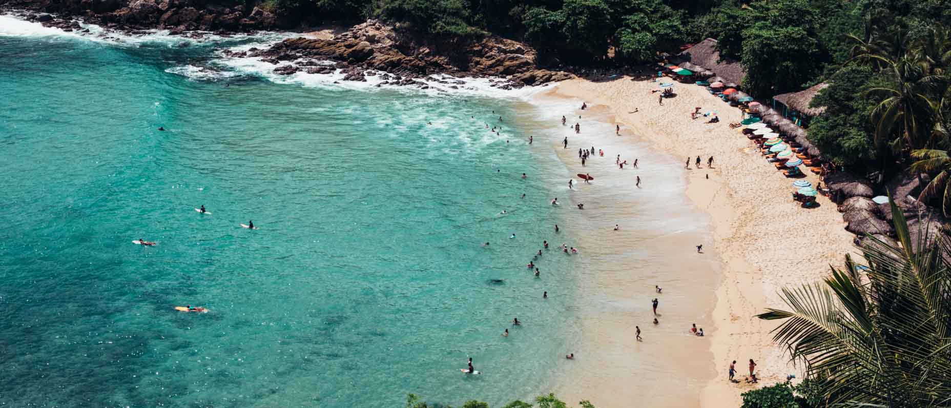 An overhead view of a sandy beach surrounded by dense trees. Several people swim or sit on surfboards in the aqua water.