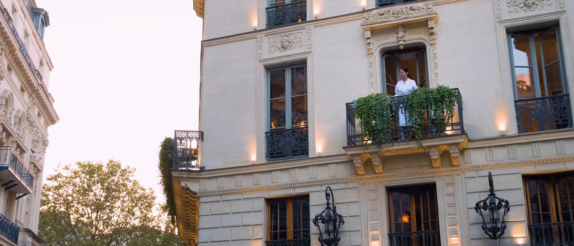 A woman in a white robe stands on the small balcony of an ornate, stone-faced building. Steel-framed windows and ornate ironwork are representative of classic European architecture.   