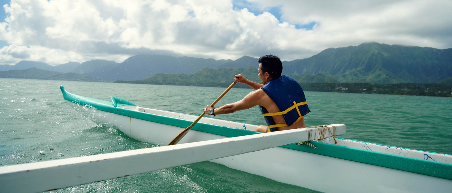 Man paddling a canoe through shimmering turquoise water, surrounded by scenic mountains, beneath a cloudy sky 