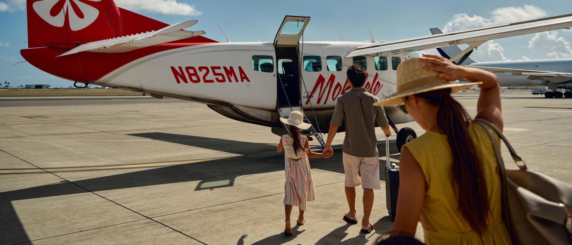 Two adults and one child walk on a tarmac towards a small red-and-white plane under a sunny sky with a few clouds.