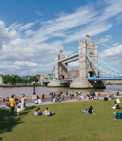 A view of Tower Bridge, London, from the South Bank. People lounge on the grass. The sky is cloudy. 