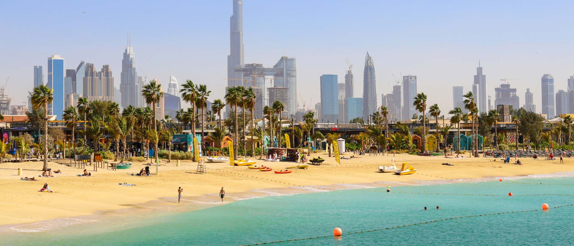 Sandstrand mit Menschen, ruhiges türkisblaues Wasser und eine Skyline mit Wolkenkratzern und Palmen im Hintergrund