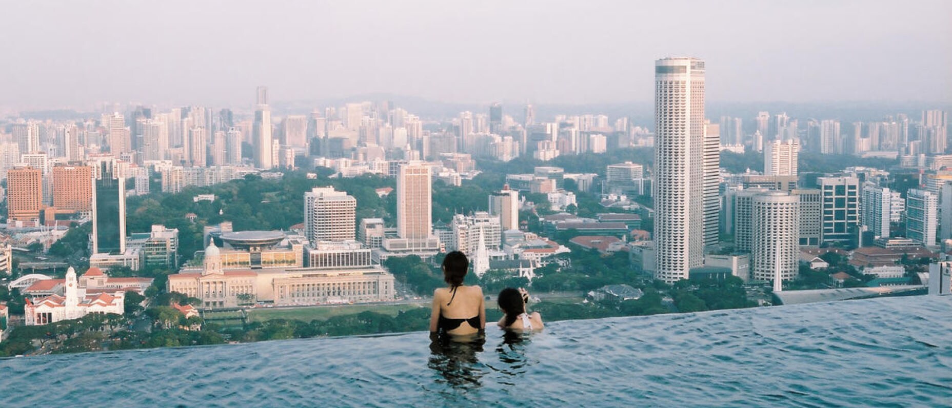 Two friends look out over a vast city skyline from the edge of a rooftop infinity pool.