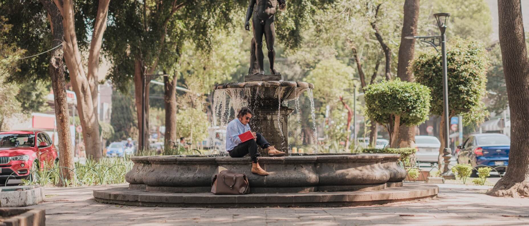 A traveler sits on the edge of a fountain writing in a notebook while a statue flows water behind them.