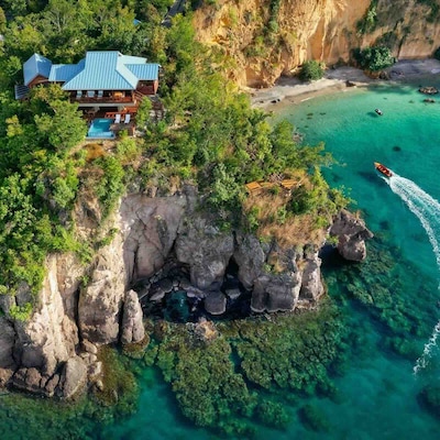Une villa isolée à flanc de colline surplombe les eaux turquoise d’une côte rocheuse, tandis qu’un bateau à moteur passe à proximité.