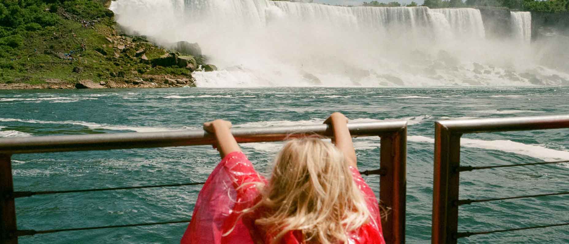 A young girl in a red poncho looks out over Niagara Falls from behind a wooden railing, with mist rising from the water. 