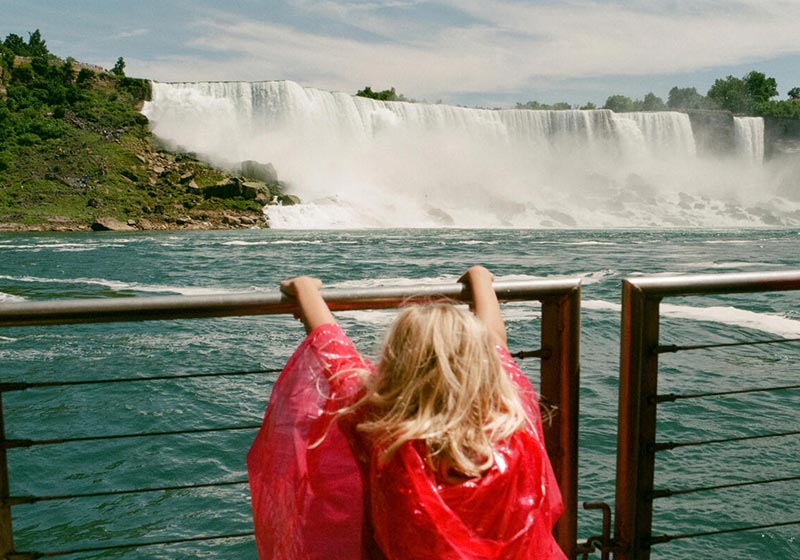 A young girl in a red poncho looks out over Niagara Falls from behind a wooden railing, with mist rising from the water. 