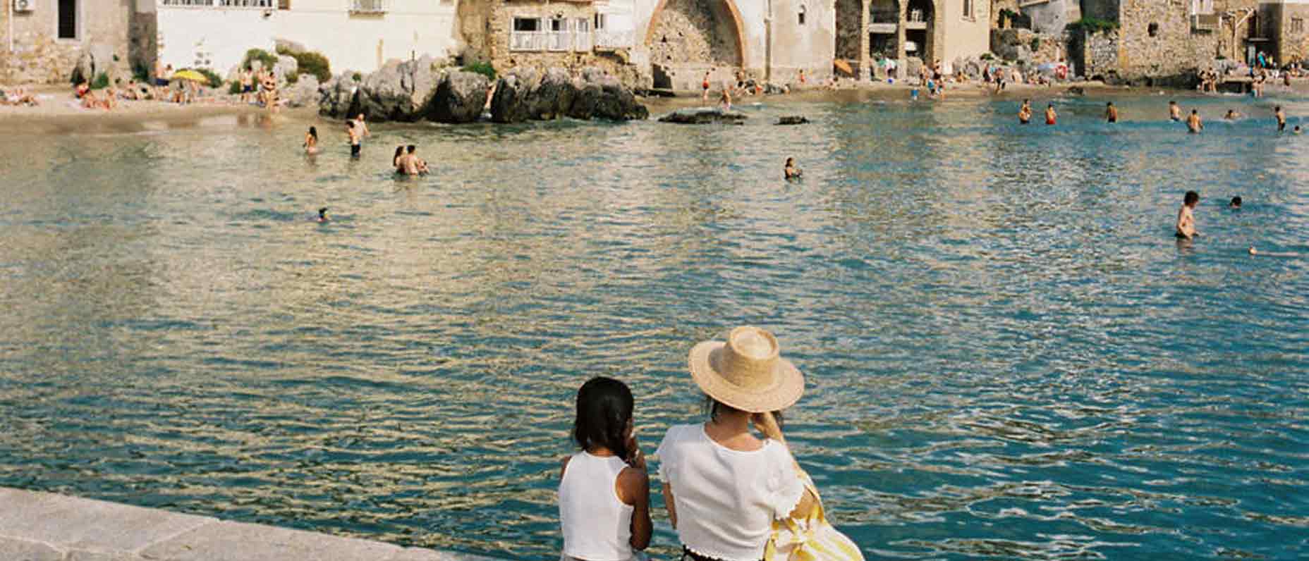 A woman and child sit by the edge of a calm coastal town, watching swimmers in the sea with historic buildings in the background.
