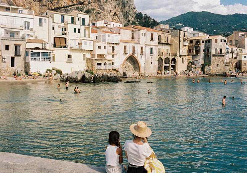 A woman and child sit by the edge of a calm coastal town, watching swimmers in the sea with historic buildings in the background.