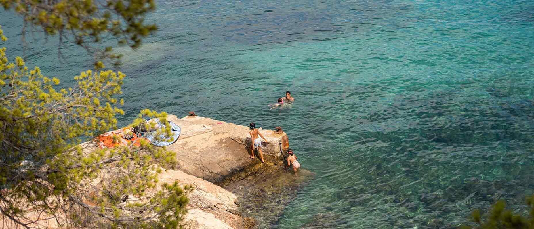 An einem sonnigen Tag entspannen sich Personen auf einem Felsen, andere spielen und schwimmen im blauen Meer.