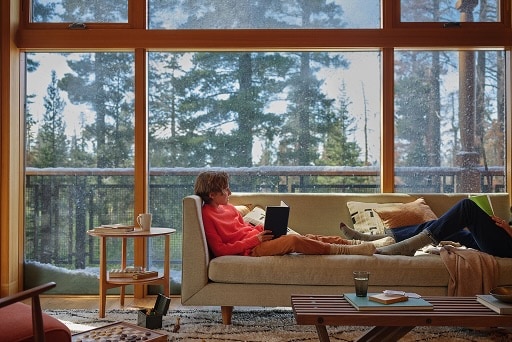 Une personne se détend en lisant un livre sur un canapé. La fenêtre en arrière-plan donne sur de grands arbres.