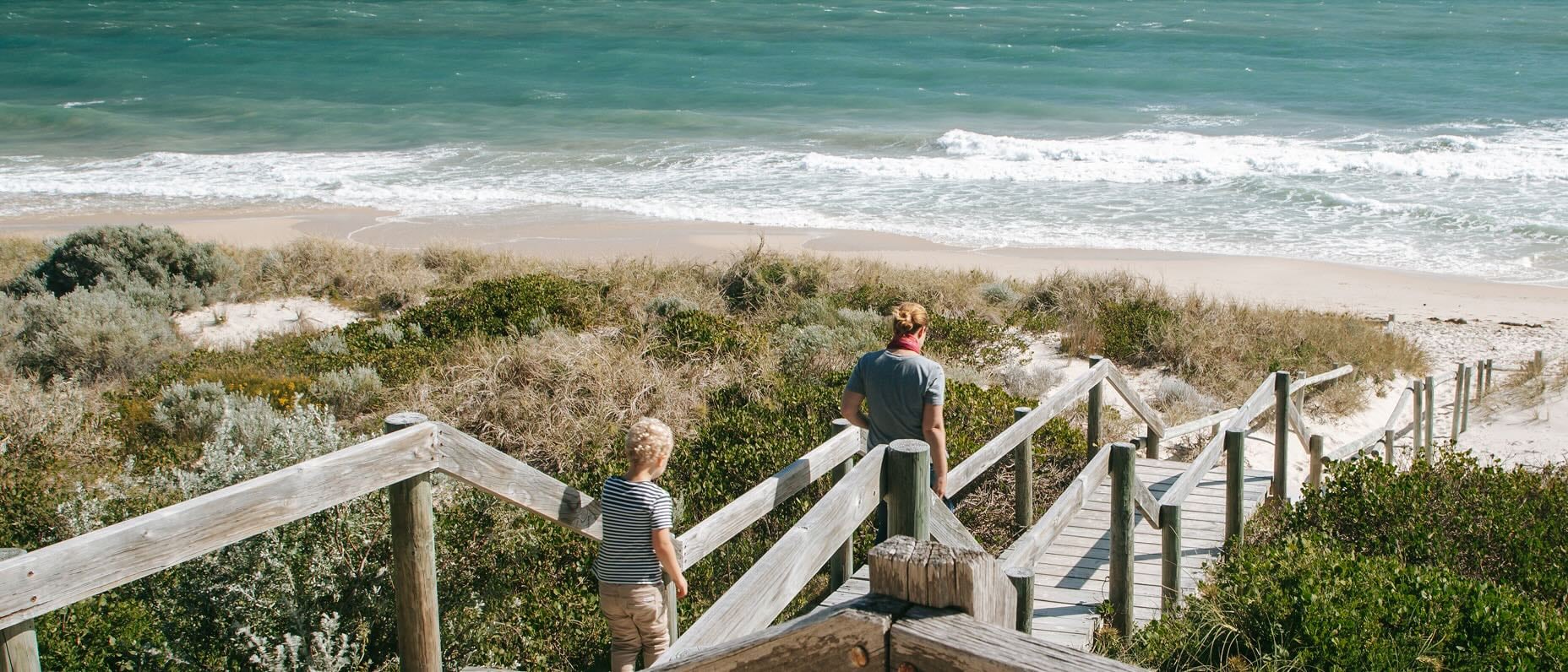 A toddler and their parent walk down wooden steps on their way to the beach.