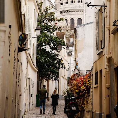 Un couple se promène main dans la main dans une ruelle étroite, entourée de murs de briques et baignée par la douce lumière de l’après-midi.