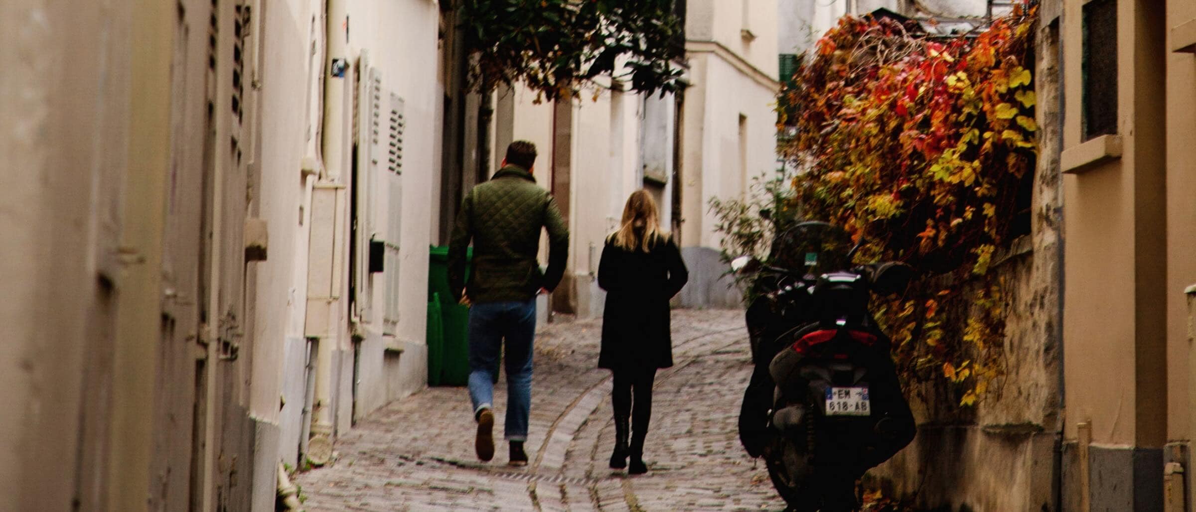 Un couple se promène main dans la main dans une ruelle bordée de murs de briques, dans la douce lumière de l’après-midi.