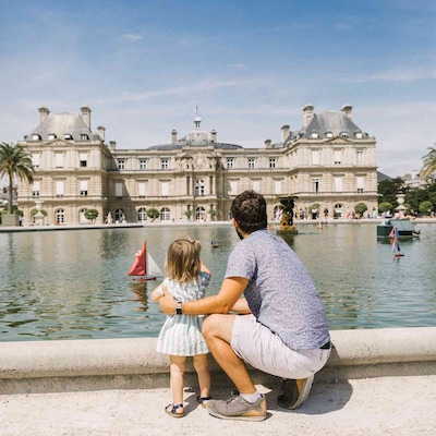A father and young daughter stand by the edge of a large fountain pond, watching toy sailboats float in the water. Behind them is the historic Luxembourg Palace in Paris, France, under a bright blue sky.