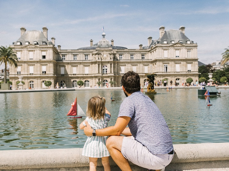 A father and young daughter stand by the edge of a large fountain pond, watching toy sailboats float in the water. Behind them is the historic Luxembourg Palace in Paris, France, under a bright blue sky.