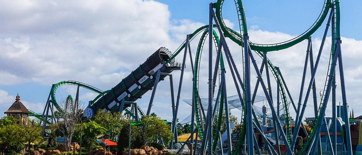 A group of people hang upside down on a green and purple roller coaster against a blue sky, hands raised in excitement.