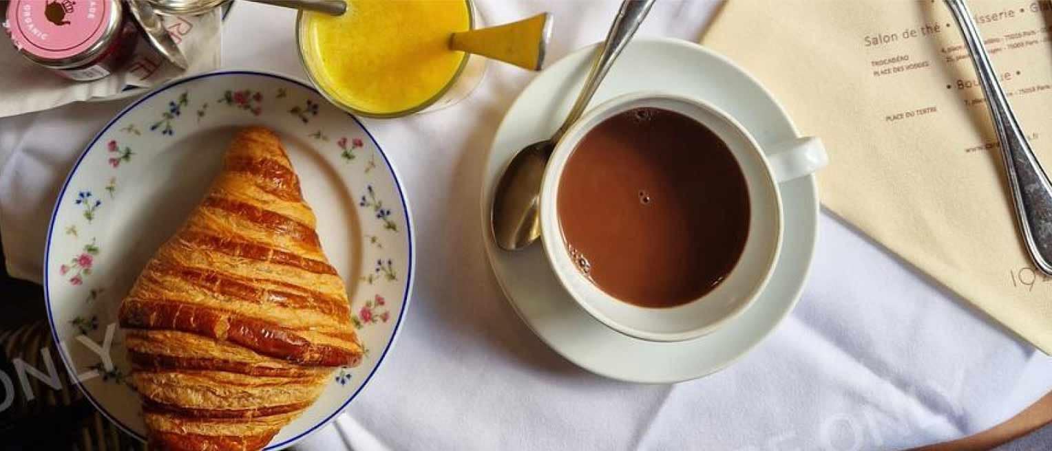 A croissant on a floral plate, with hot chocolate, orange juice, and a menu on a white tablecloth.
