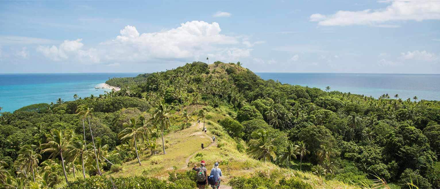 Hikers walk along a narrow, scenic ridge surrounded by palm trees and ocean on both sides, under a bright blue sky.