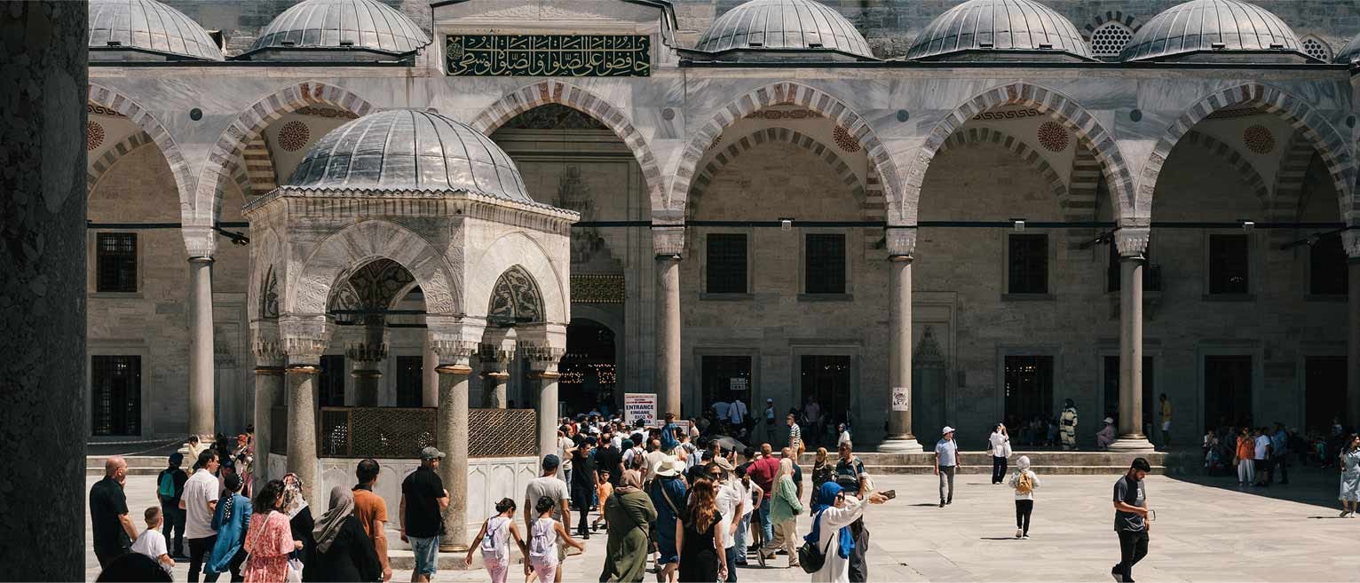 Tourists explore the grand courtyard of the Blue Mosque in Istanbul, with domed roofs, stone arches, and intricate Islamic architecture.